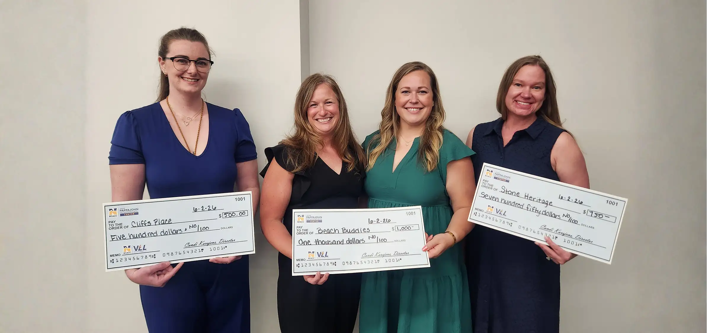 Four people smiling at the camera, holding large checks that they won from the VLL pitch competition