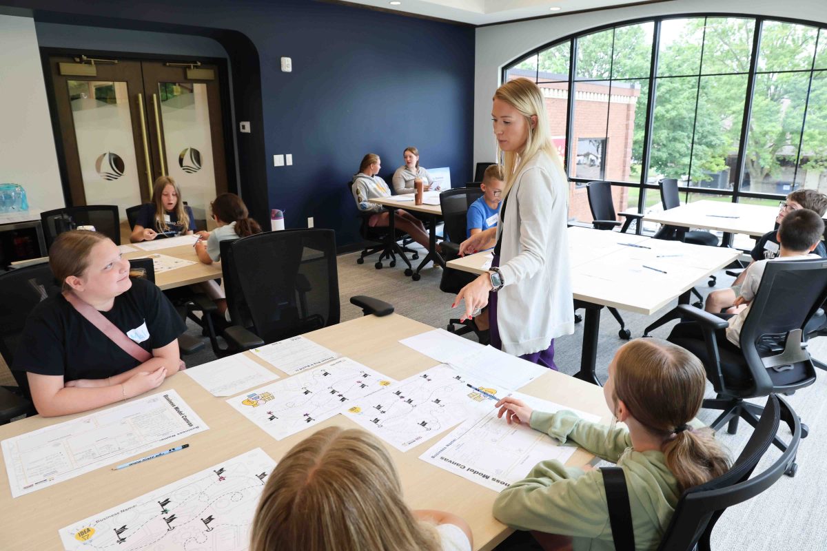 A female teacher standing in the middle of a room, talking. There are five tables with multiple male and female middle school students seated at them.