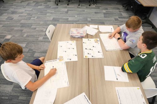 Three middle school students sitting at a large table writing on pieces of paper.