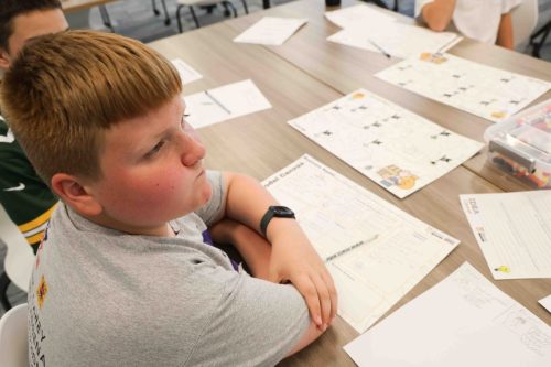 A male middle school student sitting at a desk with multiple papers and worksheets scattered across it.