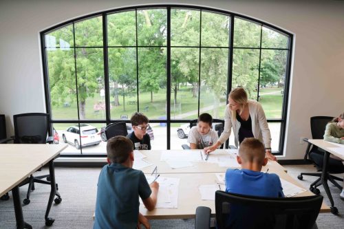 A female teacher standing at a table where four male middle school students are seated. The teacher is leaning over to help a student with his work.