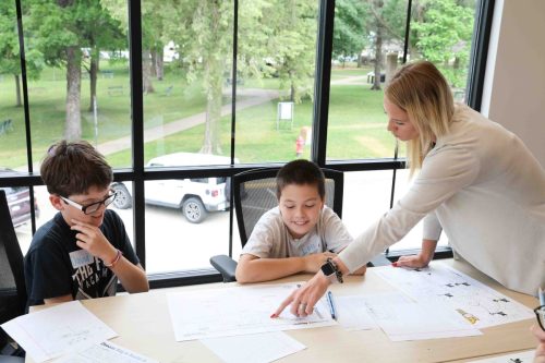 A female teacher leaning on a table talking to two male middle school students who are seated working at the same table. 